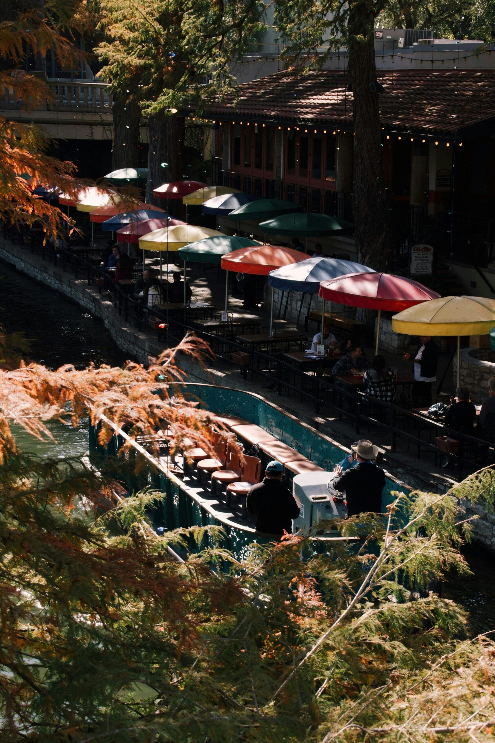Vibrant umbrellas line a riverside café, with tourists enjoying a boat ride in this scenic urban landscape.